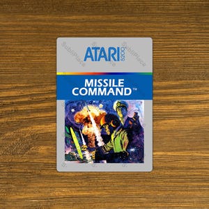 May include: A vintage Atari 5200 game cartridge for Missile Command. The cartridge features a colorful label with the game's title and artwork depicting a missile launch and military figures. The background is a light gray.