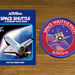 May include: A blue and red Space Shuttle Pilot patch with a white space shuttle flying over a rainbow and stars. The patch has the text "Space Shuttle Pilot" in white letters on a blue background and "ACTIVISION" in white letters on a red background.