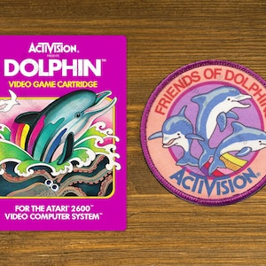 May include: A pink and purple video game cartridge for the Atari 2600 video game system. The cartridge features a dolphin jumping out of the water with a rainbow-colored tail. The text on the cartridge reads "ACTIVISION PRESENTS DOLPHIN™ VIDEO GAME CARTRIDGE FOR THE ATARI 2600™ VIDEO COMPUTER SYSTEM™".