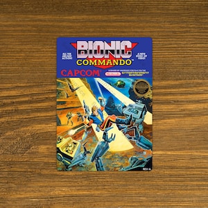 May include: Bionic Commando video game box art. The cover features a futuristic scene with a hero in blue and orange, robots, and the Capcom logo. The Nintendo Entertainment System logo is also visible.