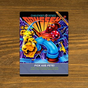 May include: A video game cartridge for the Odyssey 2 console. The cartridge is blue and yellow with a cartoon image of a miner holding a pickaxe. The text on the cartridge reads "CHALLENGER SERIES EXPANDED MEMORY!! PICK AXE PETE! Thrilts, chillis and spills as Pick Axe Pete strikes it rich is the Misty Mountain Mine!"