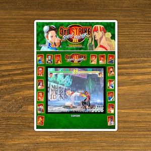 May include: A green and white arcade cabinet screen showing a fighting game with two characters, one in red and one in white, fighting in a rainy street. The game is called "3rd Strike Street Fighter III: New Edition". The screen also shows a character selection screen with a variety of characters to choose from.
