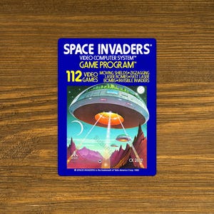 May include: A blue and white Atari game cartridge for Space Invaders. The cartridge has a picture of a spaceship and the text "Space Invaders Video Computer System™ Game Program™ 112 Video Games Moving Shields Zigzagging Laser Bombs Fast Laser Bombs Invisible Invaders CX 2632".