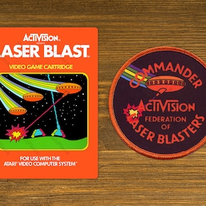 May include: An orange rectangular Activision Laser Blast video game cartridge box featuring a space battle graphic, alongside a circular embroidered patch with a dark background, orange border, and the text Commander Activision Federation of Laser Blasters.