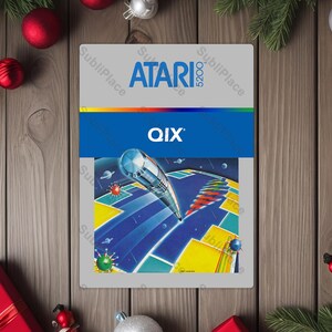 May include: A rectangular Atari 5200 game cartridge featuring the game "QIX". The cartridge has a gray background with the Atari logo and game title in blue. The game art depicts a futuristic scene with geometric shapes.