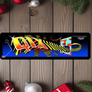 May include: A black and blue arcade game cabinet marquee with the word "Qix" in yellow, red, and black letters. The marquee also features a black and yellow striped shape and a blue and white geometric pattern. The text "TAITO AMERICA CORPORATION © 1981" is printed in white on the marquee.