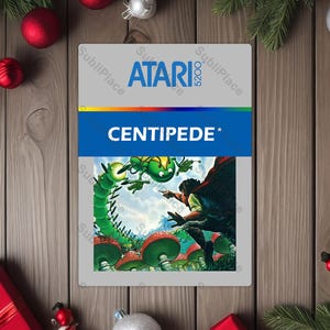 May include: Atari 5200 Centipede game cartridge. The cartridge features the Atari logo and the game title in white text on a blue background. The game's artwork depicts a green centipede and a figure in a red cape. The cartridge is surrounded by Christmas decorations.