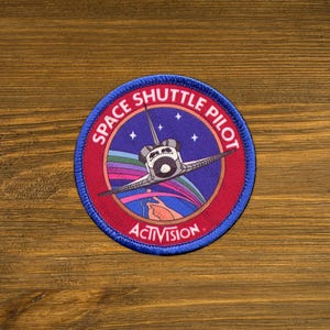 May include: A red and blue embroidered patch with a space shuttle flying over a rainbow and stars. The text "SPACE SHUTTLE PILOT" is written in a circle around the shuttle. The text "ACTIVISION" is written below the shuttle.