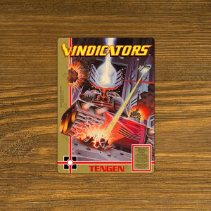 May include: A vintage Vindicators arcade game cartridge. The artwork features a futuristic battle scene with a red vehicle firing a laser beam. The game title is in yellow, and the Tengen logo is visible.