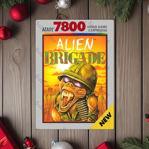 May include: Atari 7800 Alien Brigade video game cartridge. The cover art features a menacing alien soldier with a helmet and open mouth. The background is a sunset scene with the word "NEW" on a yellow corner.