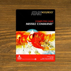 May include: A vintage Atari Missile Command game box. The box features a black top with white text reading "ATARI 400/800" and "COMPUTER GAME MISSILE COMMAND". The bottom half displays a colorful illustration of a missile defense scene.
