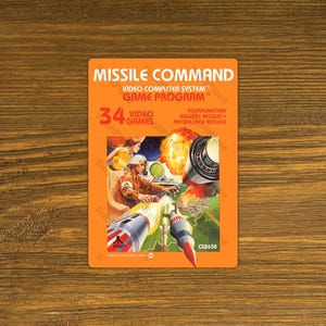 May include: An orange Atari Missile Command video game cartridge. The game program features a graphic of a pilot in a cockpit with explosions and missiles. The cartridge includes the text "34 Video Games" and "Video Computer System Game Program".