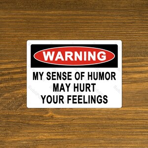 May include: White warning sign with black and red text. The sign reads "WARNING MY SENSE OF HUMOR MAY HURT YOUR FEELINGS".