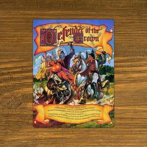 May include: A rectangular card featuring the title "Defender of the Crown" with a medieval battle scene. Knights on horseback clash with swords, a woman is held, and a castle is in the background. The card is on a wooden surface.