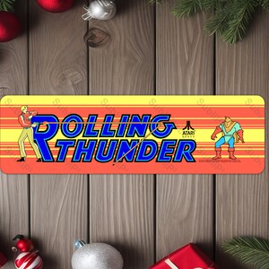 May include: A yellow and orange arcade cabinet marquee with the text "Rolling Thunder" in blue letters. The text "Atari Games" is in white letters. There are two characters on the marquee, one on the left and one on the right.