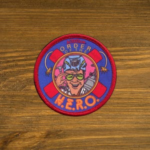 May include: A round patch with a red border and a blue background. The patch features a cartoon illustration of a person wearing a helmet and sunglasses. The text "ORDER OF THE HERO" and "N.E.R.O." are displayed.