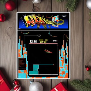 May include: A sticker featuring the QIX arcade game. The top section has the game's logo in yellow, orange, and black. The main game screen is black with colorful geometric shapes and text. The sticker is surrounded by Christmas decorations.