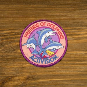 May include: A circular embroidered patch featuring three blue dolphins leaping against a light purple background. The text FRIENDS OF DOLPHINS is arched across the top, and the word ACTIVISION is printed in bold blue letters at the bottom.