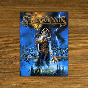 May include: A rectangular card featuring the title "Shadowlands" in gold lettering. The artwork depicts a fantasy scene with a robed figure, lightning, and a battle scene, set against a blue and orange sky. The word "DOMARK" is at the bottom.