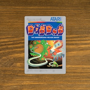 May include: A Dig Dug video game cartridge for the Atari 5200 SuperSystem. The cartridge features the game's logo in orange and blue, along with illustrations of characters from the game. The text "THE UNDERGROUND ARCADE SMASH!" is also visible.