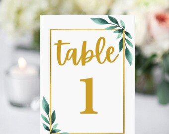 May include: White table number card with gold lettering and a gold frame. The card says "table 1" and has green watercolor leaves in the corners. The card is on a small wooden easel.