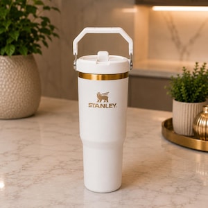May include: A white insulated tumbler with a gold-colored rim and a matching white handle on the lid. The Stanley logo, featuring a winged bear, is printed in gold on the front of the cup. The tumbler has a tapered base and a smooth matte finish.
