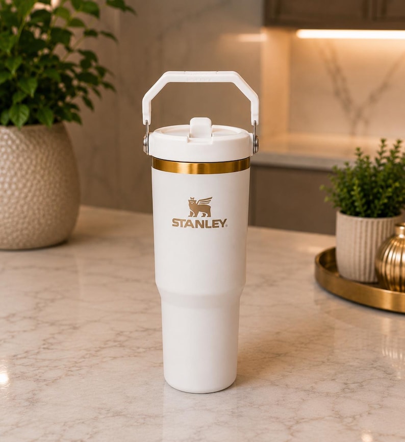 May include: A white insulated tumbler with a gold-colored rim and a matching white handle on the lid. The Stanley logo, featuring a winged bear, is printed in gold on the front of the cup. The tumbler has a tapered base and a smooth matte finish.