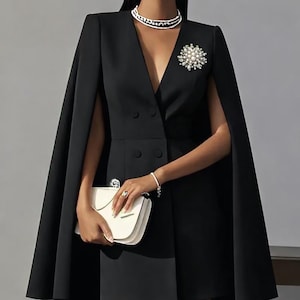 May include: A black cape dress with a double-breasted front and a V-neckline. The dress features a cape design, a pearl brooch, and a white clutch. A black and white necklace and a bracelet complete the look.