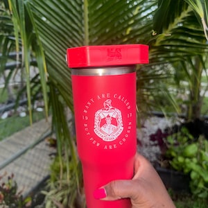 May include: A bright red insulated tumbler with a matching red lid. The front features a white circular emblem with Greek letters and the text "MANY ARE CALLED BUT FEW ARE CHOSEN" along with the numbers "19" and "13" on either side of the central crest.