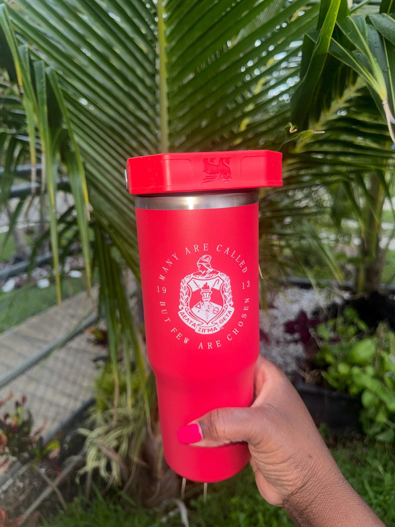 May include: A bright red insulated tumbler with a matching red lid. The front features a white circular emblem with Greek letters and the text "MANY ARE CALLED BUT FEW ARE CHOSEN" along with the numbers "19" and "13" on either side of the central crest.