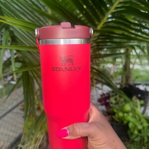 May include: A bright red Stanley tumbler with a dark red lid and a metallic silver rim. The Stanley logo, featuring a winged bear, is embossed on the front of the cylindrical vessel. The cup has a smooth, matte finish and a sturdy, functional design.
