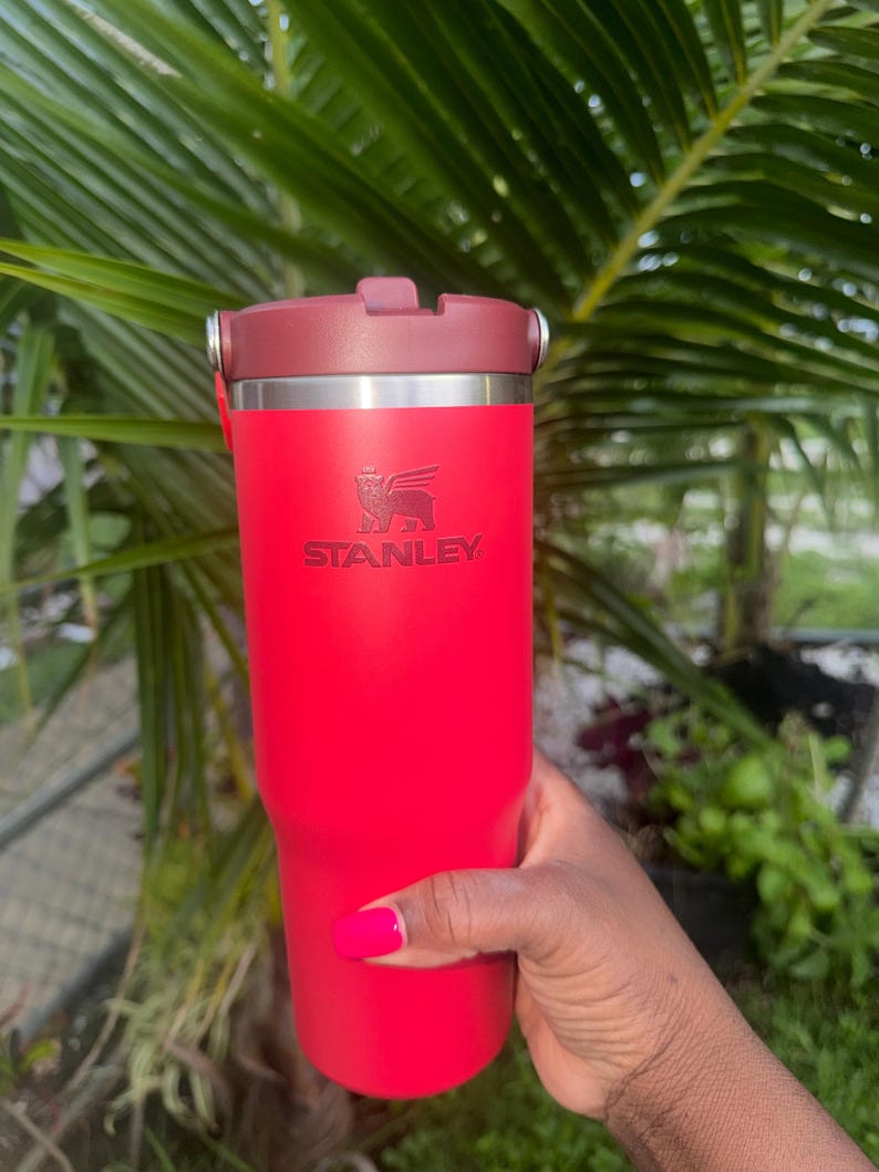 May include: A bright red Stanley tumbler with a dark red lid and a metallic silver rim. The Stanley logo, featuring a winged bear, is embossed on the front of the cylindrical vessel. The cup has a smooth, matte finish and a sturdy, functional design.