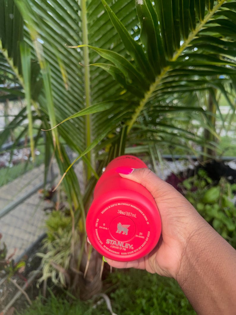 May include: A bright red Stanley brand tumbler with a 30oz capacity. The base of the tumbler features printed text including the brand logo, capacity, and manufacturing details. The tumbler is held in a hand with bright pink fingernail polish.