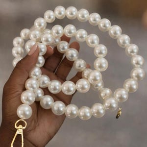 May include: A strand of large, off-white faux pearls. The pearls are strung together, forming a long necklace or strap. A gold-colored clasp and small charm are attached to the end of the strand. The pearls are held in a hand.