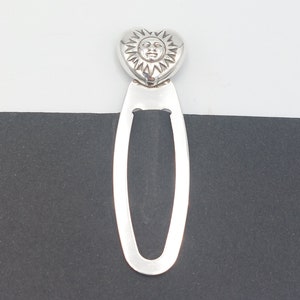 Puede incluir: Un marcapáginas plateado con una parte superior en forma de corazón con un diseño de cara de sol. El marcapáginas tiene una forma ovalada alargada, perfecto para marcar páginas en libros. El diseño es simple y elegante.