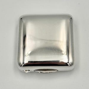 May include: A silver square compact with a hinged lid. The compact is smooth and shiny.
