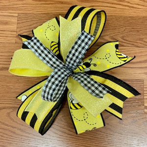 May include: A yellow and black bow with a bee pattern and black and white checkered ribbon. The bow is made of glitter fabric and has a shiny finish.