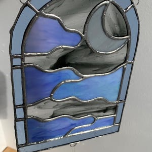 May include: A stained glass window hanging with a crescent moon and clouds design. The window is blue, purple, and gray. A crystal prism hangs from the bottom of the window.