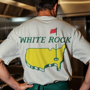 Pu&ograve; includere: Una t-shirt a maniche corte di colore chiaro con una grafica sul retro. Il design include la scritta "WHITE ROCK" in lettere verde scuro sopra una mappa gialla degli Stati Uniti con un'asta per bandiera da golf e una bandiera rossa sulla mappa.