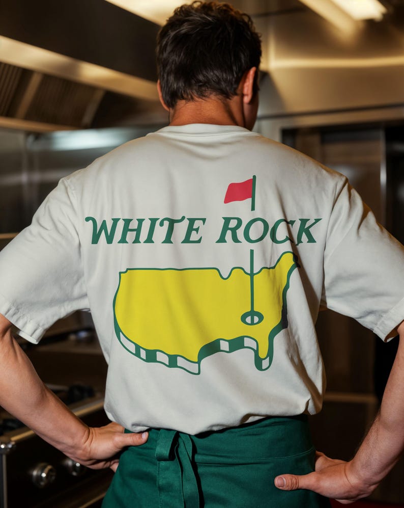 Pu&ograve; includere: Una t-shirt a maniche corte di colore chiaro con una grafica sul retro. Il design include la scritta "WHITE ROCK" in lettere verde scuro sopra una mappa gialla degli Stati Uniti con un'asta per bandiera da golf e una bandiera rossa sulla mappa.