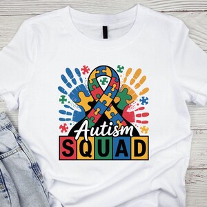 Peut inclure: T-shirt blanc avec un graphisme coloré représentant un ruban de puzzle, des empreintes de mains et les mots "Autism Squad". Le motif comprend des pièces de puzzle de différentes couleurs, symbolisant la sensibilisation à l'autisme.