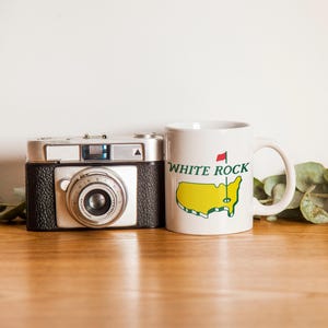 Pu&ograve; includere: Una fotocamera a pellicola in stile vintage color argento e nero accanto a una tazza in ceramica bianca. La tazza presenta una mappa gialla degli Stati Uniti, una bandierina da golf rossa e il testo verde "WHITE ROCK" stampato sul davanti.