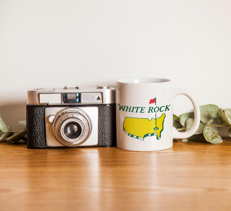 Pu&ograve; includere: Una fotocamera a pellicola in stile vintage color argento e nero accanto a una tazza in ceramica bianca. La tazza presenta una mappa gialla degli Stati Uniti, una bandierina da golf rossa e il testo verde "WHITE ROCK" stampato sul davanti.