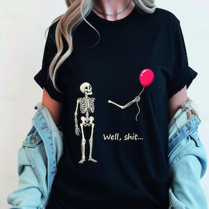 May include: A black t-shirt featuring a graphic of a skeleton standing next to a detached skeleton arm holding a red balloon. The text "Well, shit..." is printed in white below the arm. The shirt is worn with a light blue denim jacket.