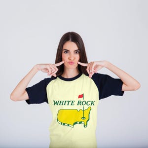 Pu&ograve; includere: Una t-shirt raglan con corpo giallo pallido e maniche blu navy scuro. La parte anteriore presenta una grafica della mappa degli Stati Uniti in giallo con un contorno verde, una bandierina da golf verde e il testo WHITE ROCK in lettere verdi.