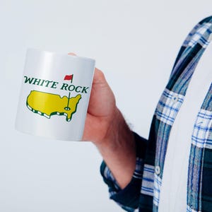 Pu&ograve; includere: Una tazza in ceramica bianca con la scritta WHITE ROCK in lettere verde scuro sopra un grafico giallo degli Stati Uniti contigui. Una piccola bandiera rossa su un'asta &egrave; posizionata sopra la mappa, simile a un segnaposto per buca da golf.