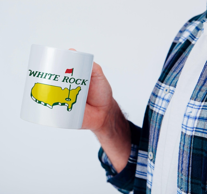 Pu&ograve; includere: Una tazza in ceramica bianca con la scritta WHITE ROCK in lettere verde scuro sopra un grafico giallo degli Stati Uniti contigui. Una piccola bandiera rossa su un'asta &egrave; posizionata sopra la mappa, simile a un segnaposto per buca da golf.
