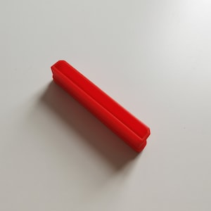 May include: A bright red, rectangular plastic object with a U-shaped channel. The object has a smooth surface and is angled slightly. The item is likely a component or part of a larger product.