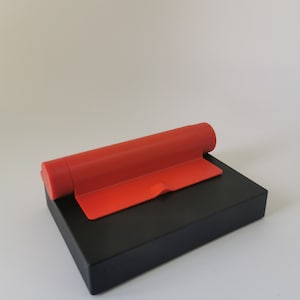 May include: A black phone stand with a red, foldable, adjustable stand. The stand is made of plastic and has a smooth, matte finish.