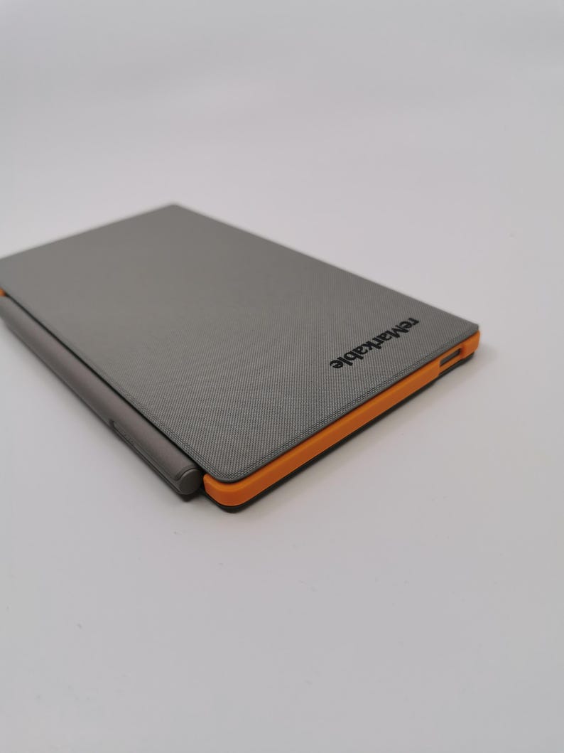 May include: A gray tablet with an orange border and a stylus. The tablet has the word "remarkable" printed on the top right corner. The tablet is closed and appears to be made of a textured material.